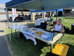 Lady sitting behind a table displaying NARFE underneath a shaded canopy.