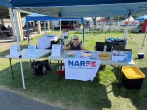 Lady sits behind table displaying NARFE underneath shaded canopy.