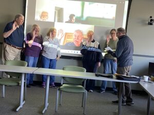 Bruce Miles, Federation Treasurer has chapter officers raise their hand to install them for 2024-2025 at the front of the meeting room. One officer, in middle of picture, was actually installed over zoom while he was at home.