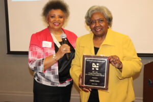 two women, the one on the right holding a plaque, the one on the left holding a microphone. Hip length and above.