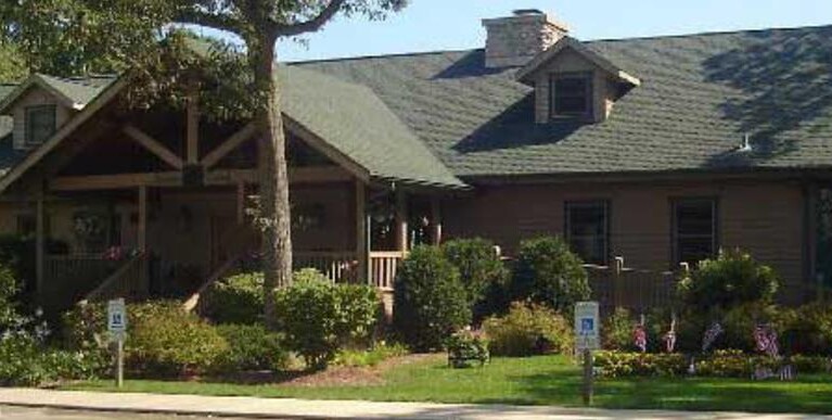 wooden log building with covered entrance, surrounded by bushes and tall trees at the front and sides