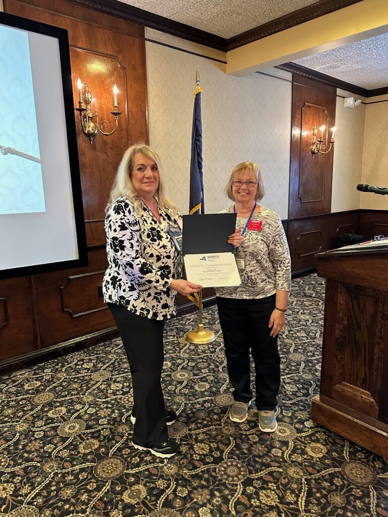 Women presenting award to another standing next to podium in front of flag.