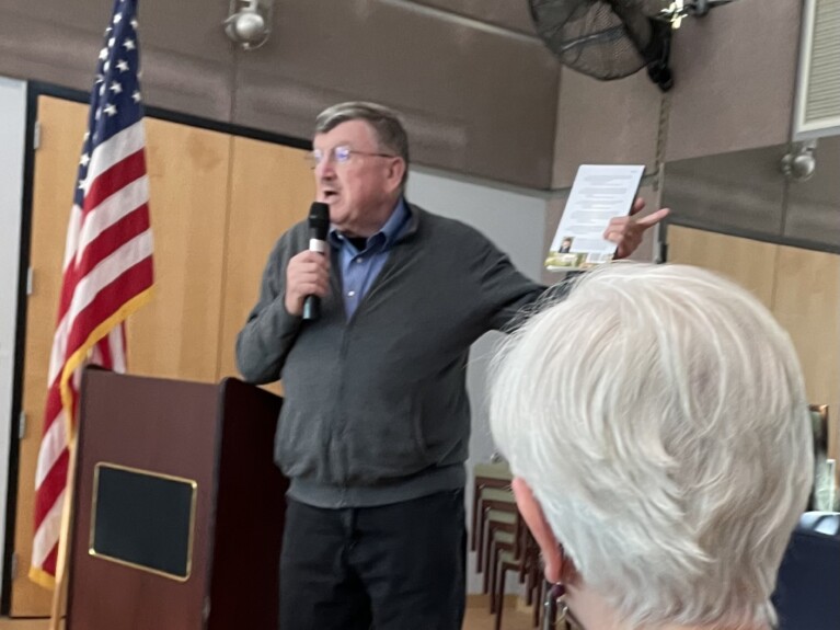 Len Lazarick by flag and podium with the back of a woman's head in the foreground