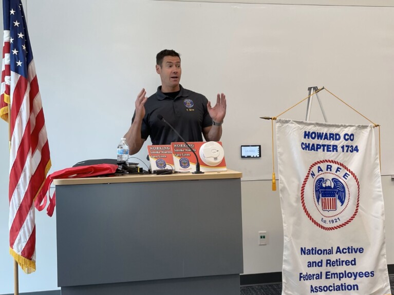 American flag, man speaking at podium with smoke alarms, a water bottle, and cloth bags, beside the Howard County NARFE Chapter 1734 banner
