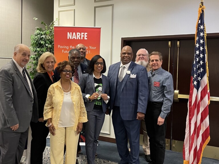 NARFE leaders presenting Angela Alsobrooks with a challenge coin in front of a NARFE banner and beside an American flag in a conference room