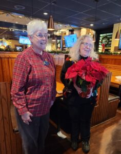 Two women in dark pants standing in a restaurant one with a poinsettia and the other in a red plaid shirt