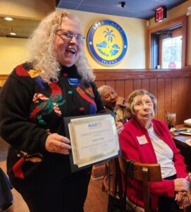 Seated woman receiving NARFE certificate from standing woman in Christmas sweater at a holiday lunch