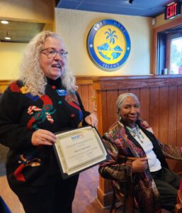 Seated woman receiving NARFE certificate from standing woman in Christmas sweater