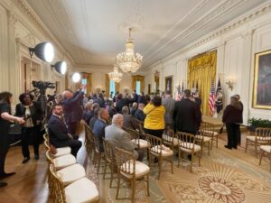 White House East Room with chandeliers, TV cameras, people sitting in rows of chairs and taking cell phone photos, with the U.S. and Presidential Seal flags lining the stage.