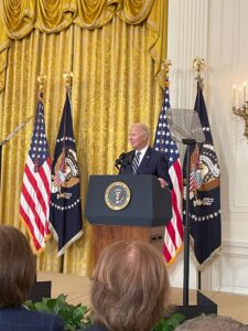 President Joe Biden speaking at a podium with the Presidential Seal in front of American and Presidential flags in the White House East Room before signing the Social Security Fairness Act.
