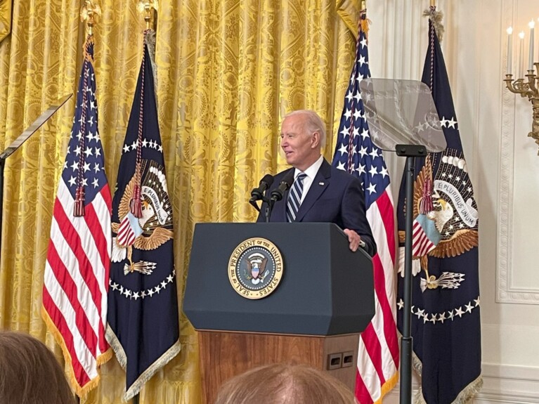 President Joe Biden speaking at a podium with the Presidential Seal in front of American and Presidential flags in the White House East Room before signing the Social Security Fairness Act.