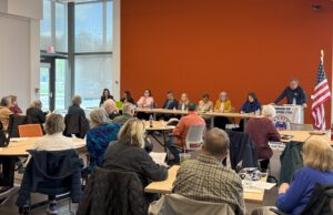 Long panel of 9 speakers in front of a bright orange wall of a large conference room with many people sitting in the audience at round tables.