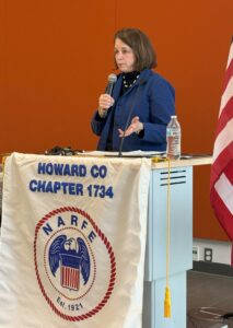Woman at a podium with microphone draped with Howard County Chapter 1734