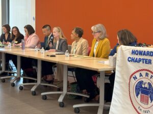 Long table of panelists beside a podium with the NARFE Howard County 1734 banner in front of an orange wall.