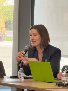 Woman seated at a table next to an open laptop, speaking into microphone in front of a full-length window.