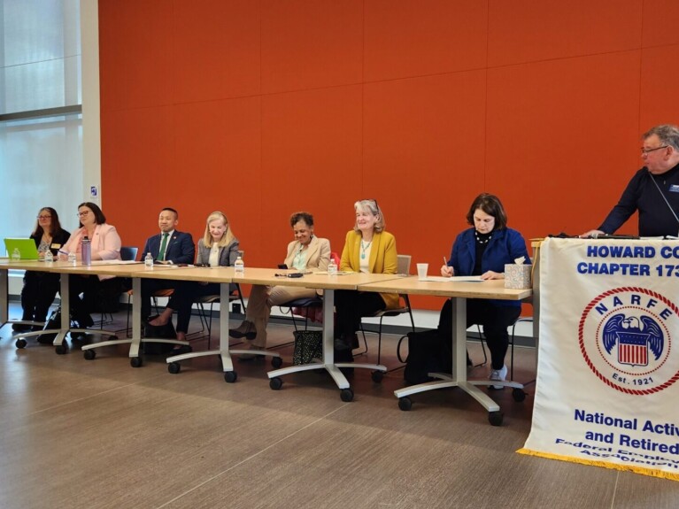 Long panel of speakers in front of a bright orange wall of a large conference room with full-length windows in the corner and a podium with a Howard County NARFE Chapter 1734 banner