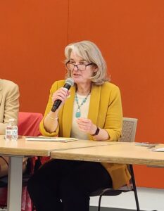 Seated woman with microphone at table in front of orange wall
