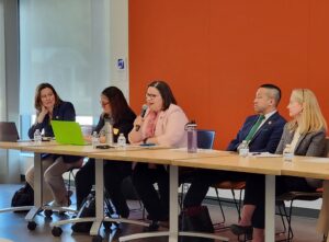 Panelists seated at a long table before an orange wall in a large conference room by a floor to ceiling window.