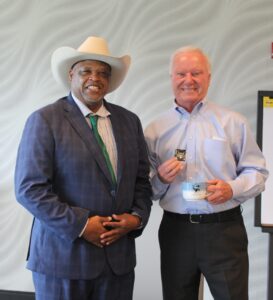 Black man in white cowboy hat and business suit and white man in blue dress shirt holding coffee mug and coin