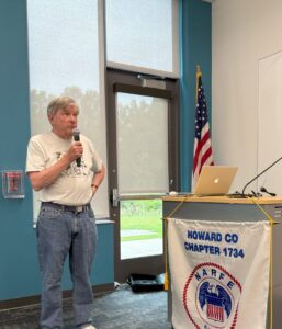 man in bird club tee shirt and jeans in front of a window and door by a podium with a NARFE Howard County banner beside an American flag
