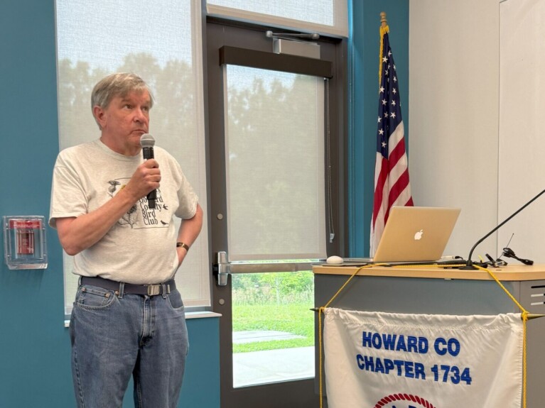 man in bird club tee shirt and jeans in front of a window and door by a podium with a NARFE Howard County banner beside an American flag