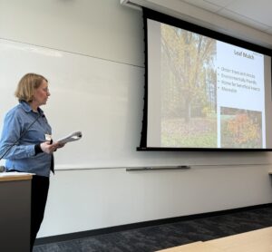 woman at podium speaking beside projected slide with pictures of trees with autumn leaves