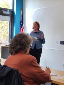 Woman by American flag and white board lecturing a woman seated at a table