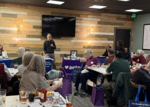 Man with microphone speaking at a table in front of a dining room of peole