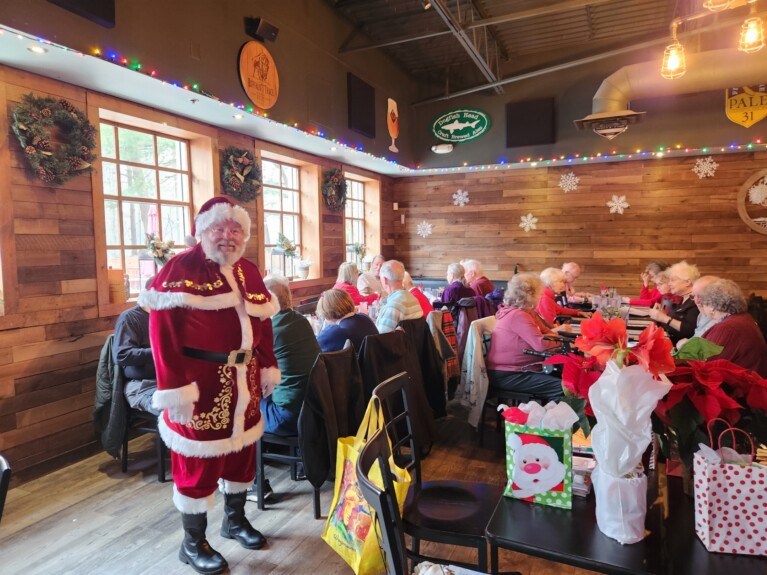 Santa Claus stands by gift laden table in a dining room with two other long tables of party-goers!