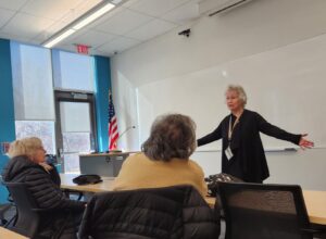 three women in a classroom. two seated facing the third who has her arms stretched out. behind her is a whiteboard, American flag and podium. in the corner is a glass exterior exit door.