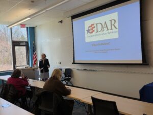 Woman beside podium, American flag, and windows in front of a classroom overhead screen