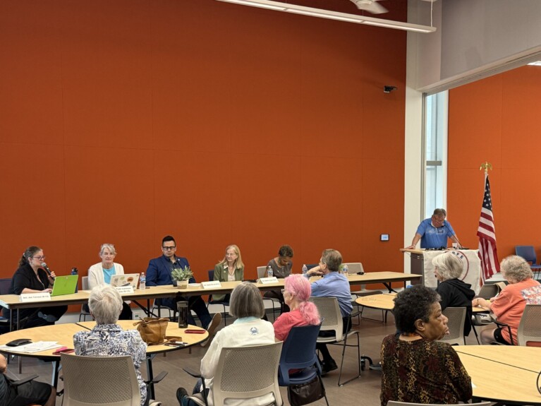 long table with panelists and a podium in front of an audience at round tables