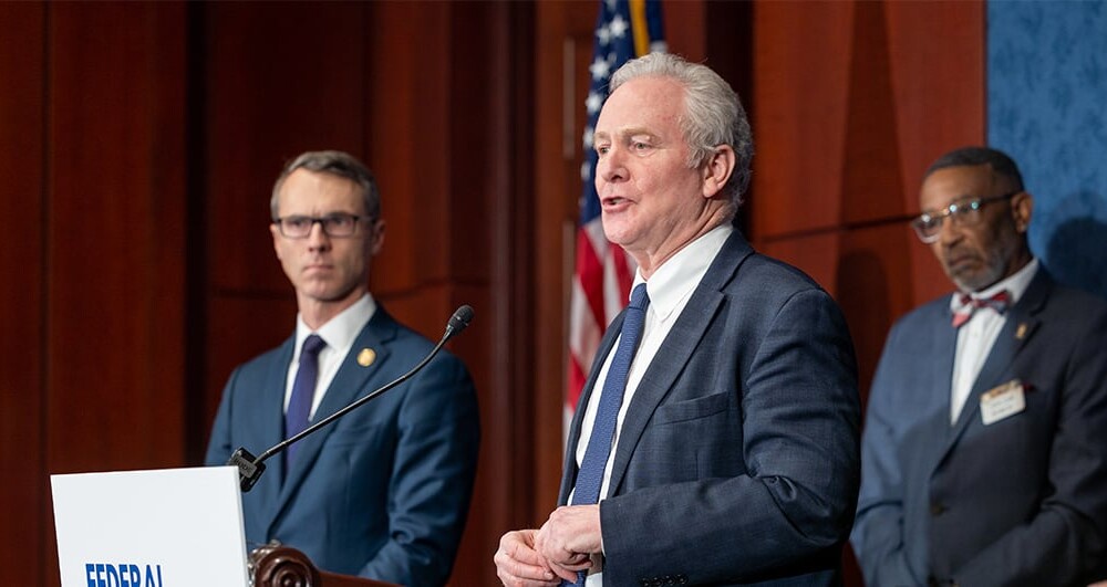 Two U.S. Legislators in suits standing at podium addressing the media with U.S. Flag in background