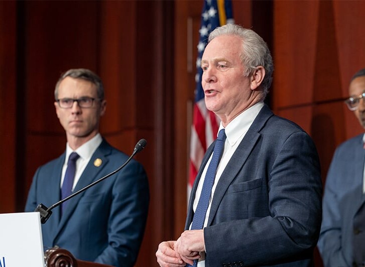 Two U.S. Legislators in suits standing at podium addressing the media with U.S. Flag in background
