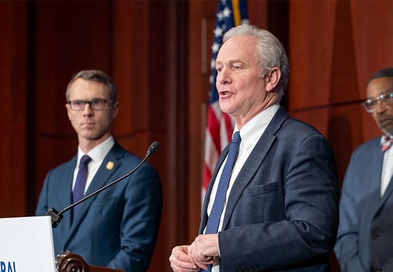 Two U.S. Legislators in suits standing at podium addressing the media with U.S. Flag in background