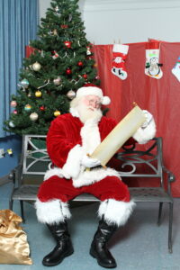 Santa is sitting in front of a Christmas Tree reading his Nice List