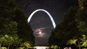 St. Louis Arch seen lite up at night between trees with moon peeking through underneath