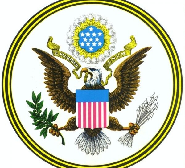 colored image of Great Seal of the United States with eagle holding banner in his beak reading E PLURIBUS UNUM, red, white & blue shield on his chest, an olive branch in his right claw and arrows in his left claw. A cloud above his head holds thirteen white stars on a blue background with a small gold circle and a larger white circle around it. The circular border has five alternating bands of black and gold colors.