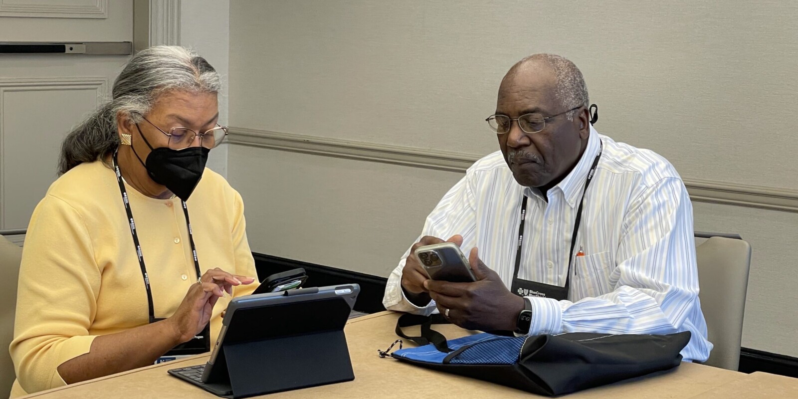 a woman with face mask and a man sit at a table with smart phone and a tablet