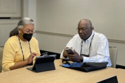 a woman with face mask and a man sit at a table with smart phone and a tablet