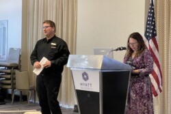 two people at front of room, a podium, and a flag