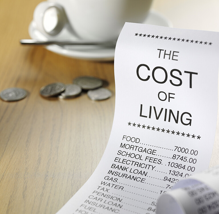 A receipt with Cost of Living at the top depicting anticipated expenses with change and a coffee cup in the background