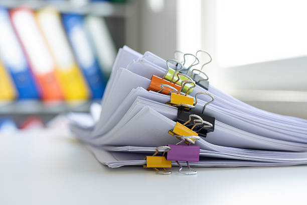 stack of papers sorted together with colorful clips and a bookcase with note binders in the background.